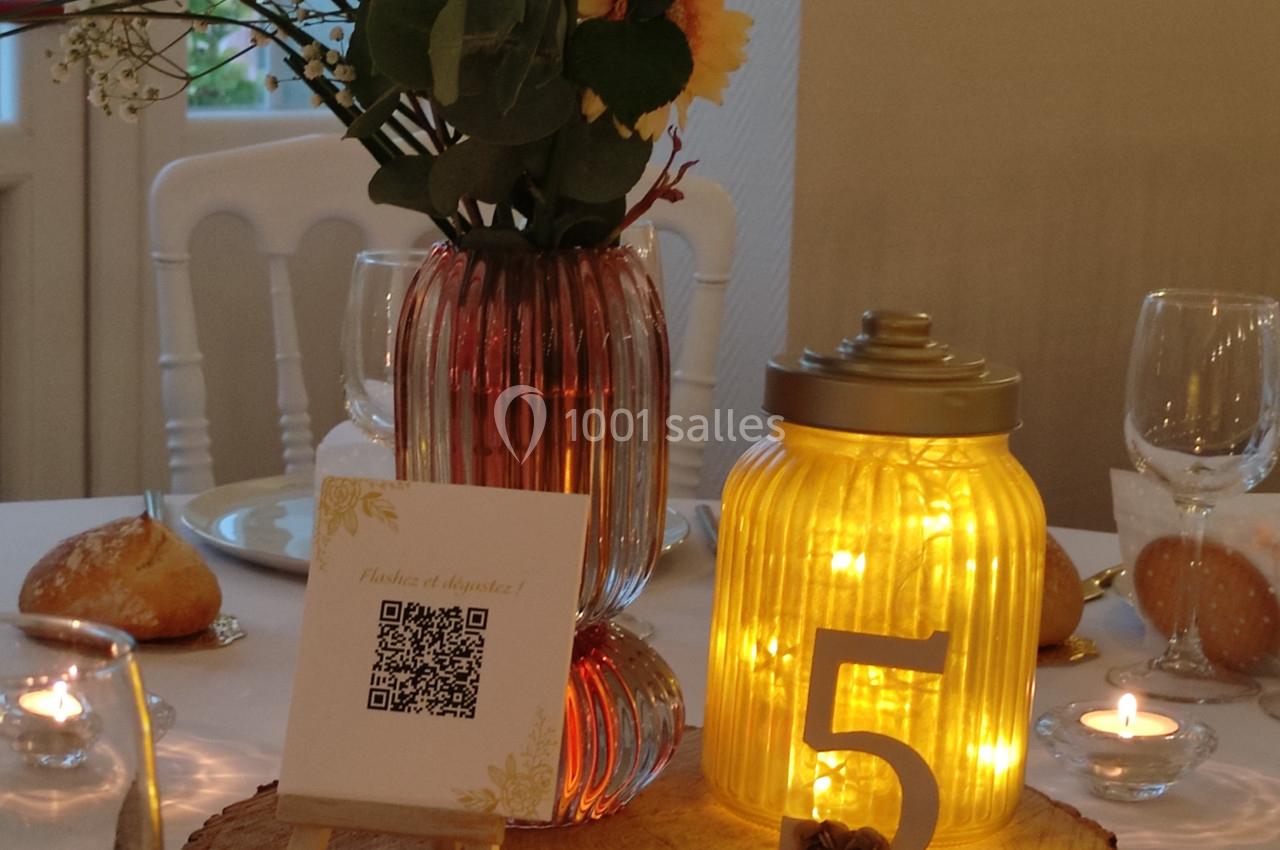 Centre de table avec fleurs, bougie LED dans un pot numéroté ’5’, QR code et lettres formant ’BEL EVENT’.