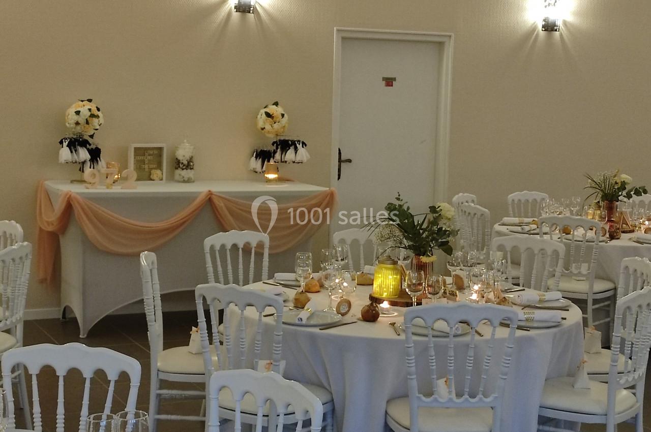 Salle de réception décorée avec des tables rondes dressées, des chaises blanches et un buffet orné de fleurs et de voilages.