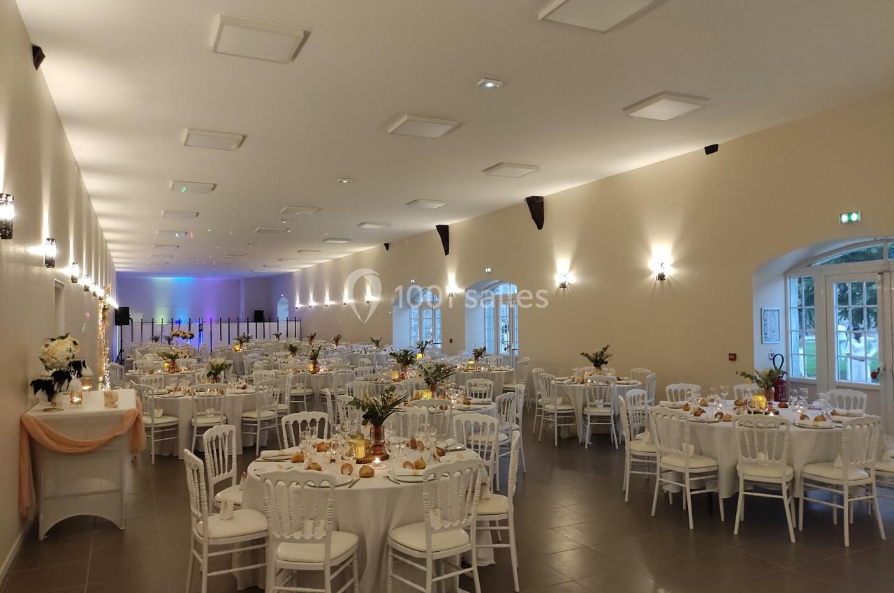 Salle de réception lumineuse avec des tables rondes décorées pour un événement, chaises blanches et éclairage chaleureux.