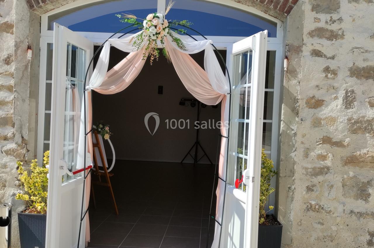 Entrée d'un bâtiment en pierre avec des portes vitrées ouvertes, décorées de voilages roses et d'une composition florale.