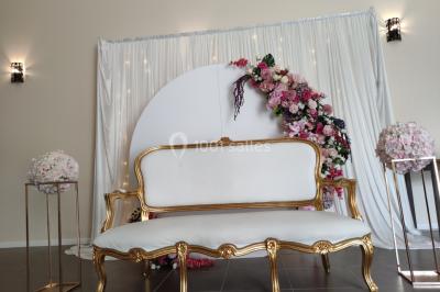 Canapé doré avec assise blanche devant un décor floral et un rideau blanc, éclairé par des appliques murales.