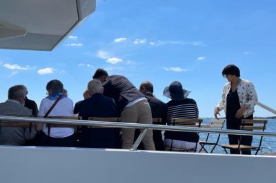 Un homme souriant assis à une table garnie de desserts, sur un bateau avec vue sur la mer et un voilier au loin.