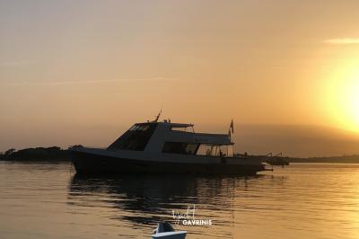 Un homme souriant assis à une table garnie de desserts, sur un bateau avec vue sur la mer et un voilier au loin.