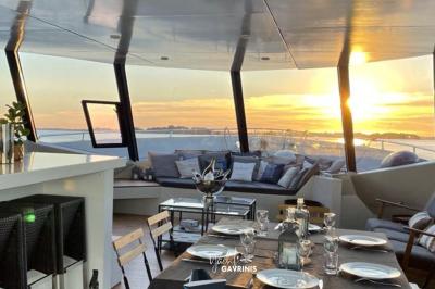 Un homme souriant assis à une table garnie de desserts, sur un bateau avec vue sur la mer et un voilier au loin.