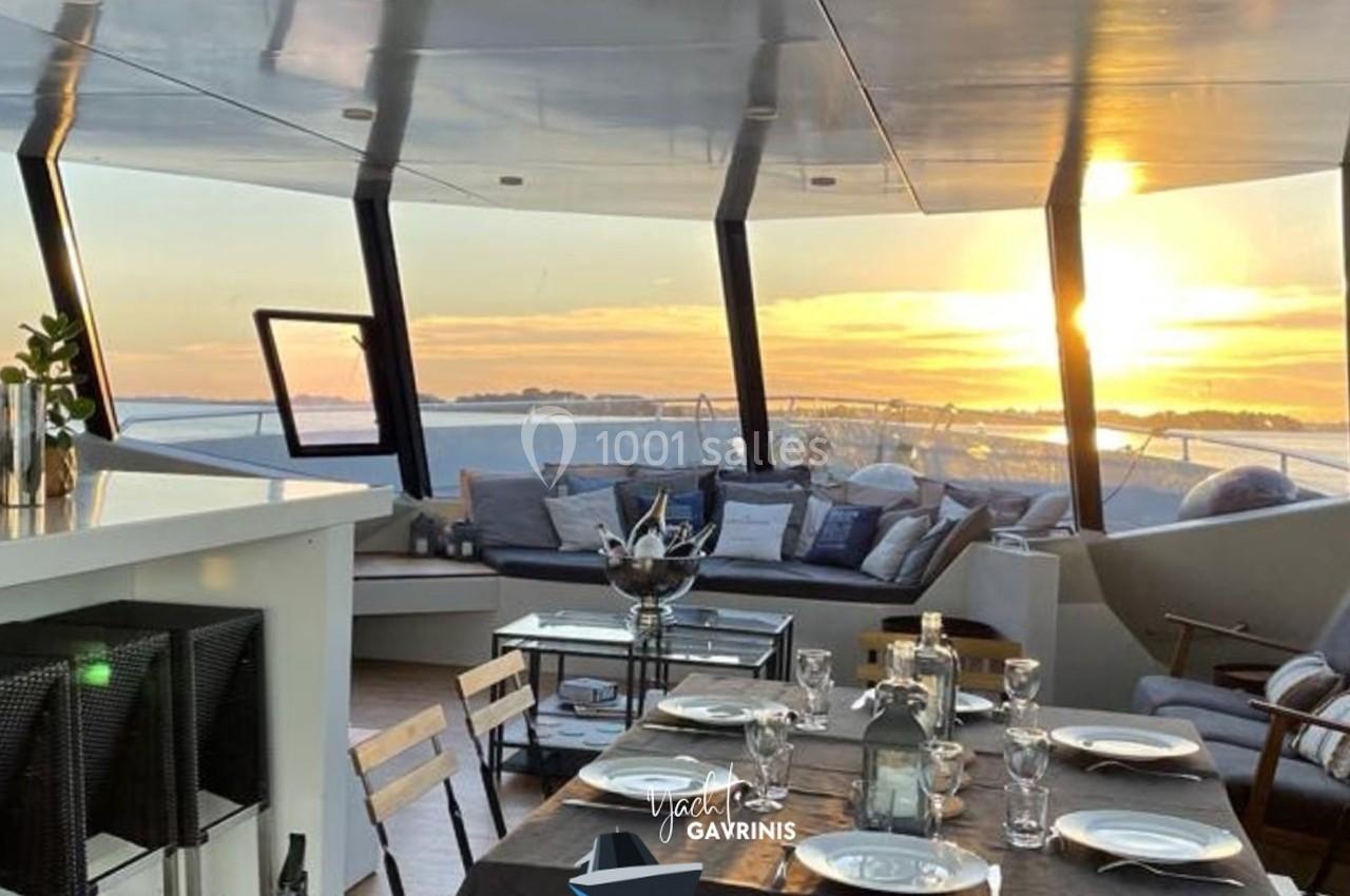 Salon extérieur d’un yacht avec table dressée, vue sur la mer et coucher de soleil à travers de grandes baies vitrées.
