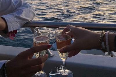 Un homme souriant assis à une table garnie de desserts, sur un bateau avec vue sur la mer et un voilier au loin.