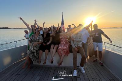 Un homme souriant assis à une table garnie de desserts, sur un bateau avec vue sur la mer et un voilier au loin.
