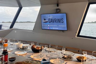 Un homme souriant assis à une table garnie de desserts, sur un bateau avec vue sur la mer et un voilier au loin.