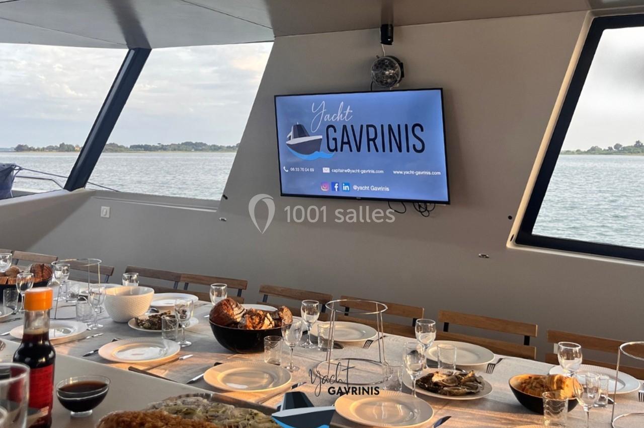Table dressée pour un repas sur un yacht avec vue sur l'eau et un écran affichant ’Yacht Gavrinis’.