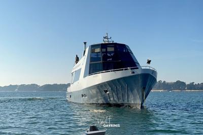 Un homme souriant assis à une table garnie de desserts, sur un bateau avec vue sur la mer et un voilier au loin.
