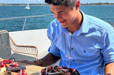 Un homme souriant assis à une table garnie de desserts, sur un bateau avec vue sur la mer et un voilier au loin.
