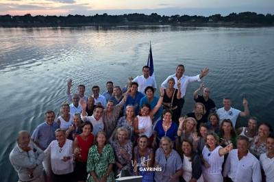 Un homme souriant assis à une table garnie de desserts, sur un bateau avec vue sur la mer et un voilier au loin.