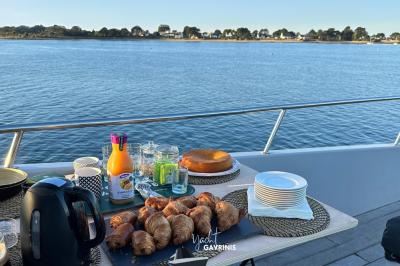 Un homme souriant assis à une table garnie de desserts, sur un bateau avec vue sur la mer et un voilier au loin.