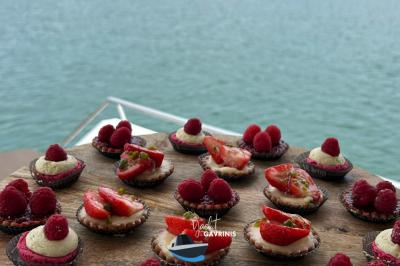 Un homme souriant assis à une table garnie de desserts, sur un bateau avec vue sur la mer et un voilier au loin.