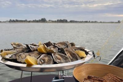 Un homme souriant assis à une table garnie de desserts, sur un bateau avec vue sur la mer et un voilier au loin.