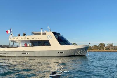 Un homme souriant assis à une table garnie de desserts, sur un bateau avec vue sur la mer et un voilier au loin.