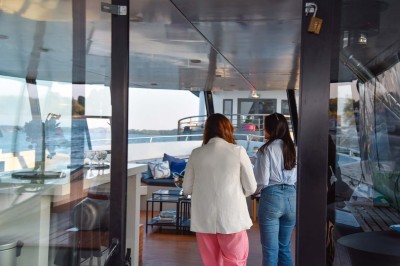Un homme souriant assis à une table garnie de desserts, sur un bateau avec vue sur la mer et un voilier au loin.