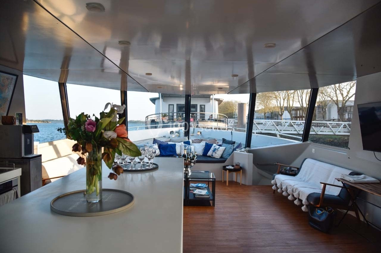Intérieur d'un bateau avec salon aménagé, table basse, canapés, fleurs sur un comptoir et vue sur l'extérieur.