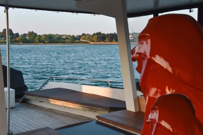Un homme souriant assis à une table garnie de desserts, sur un bateau avec vue sur la mer et un voilier au loin.