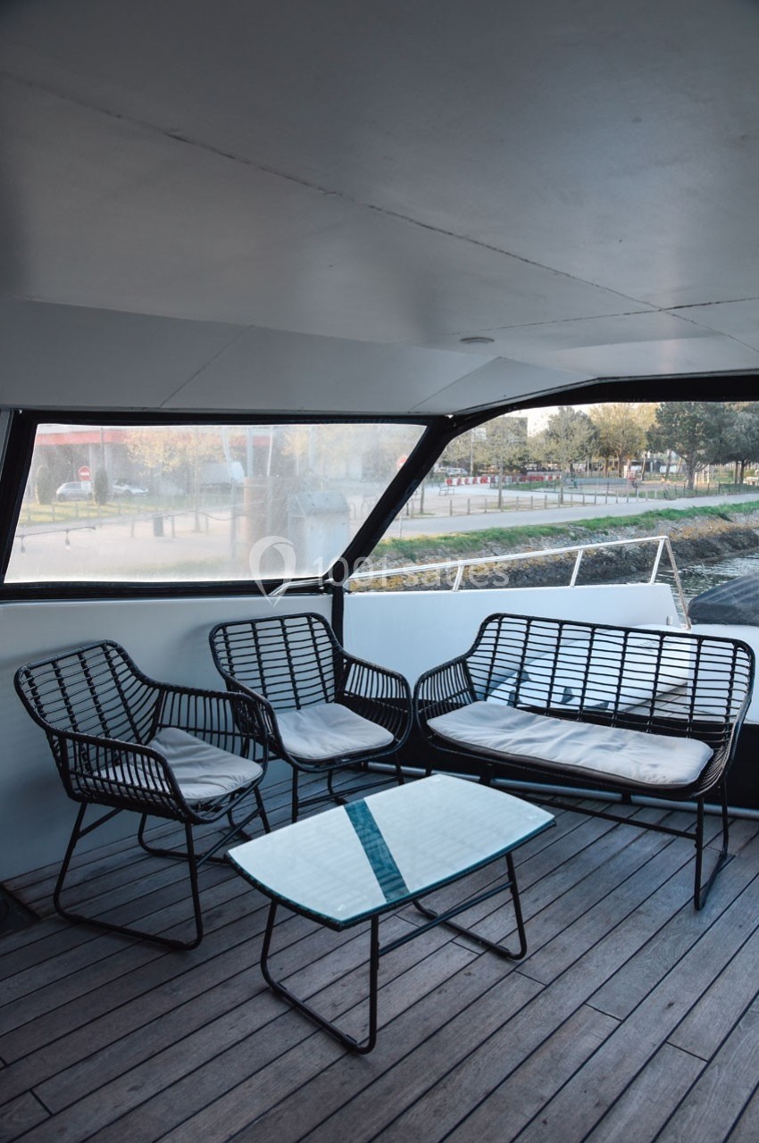 Mobilier en rotin noir avec coussins blancs sur une terrasse en bois couverte, vue sur l'extérieur urbain.