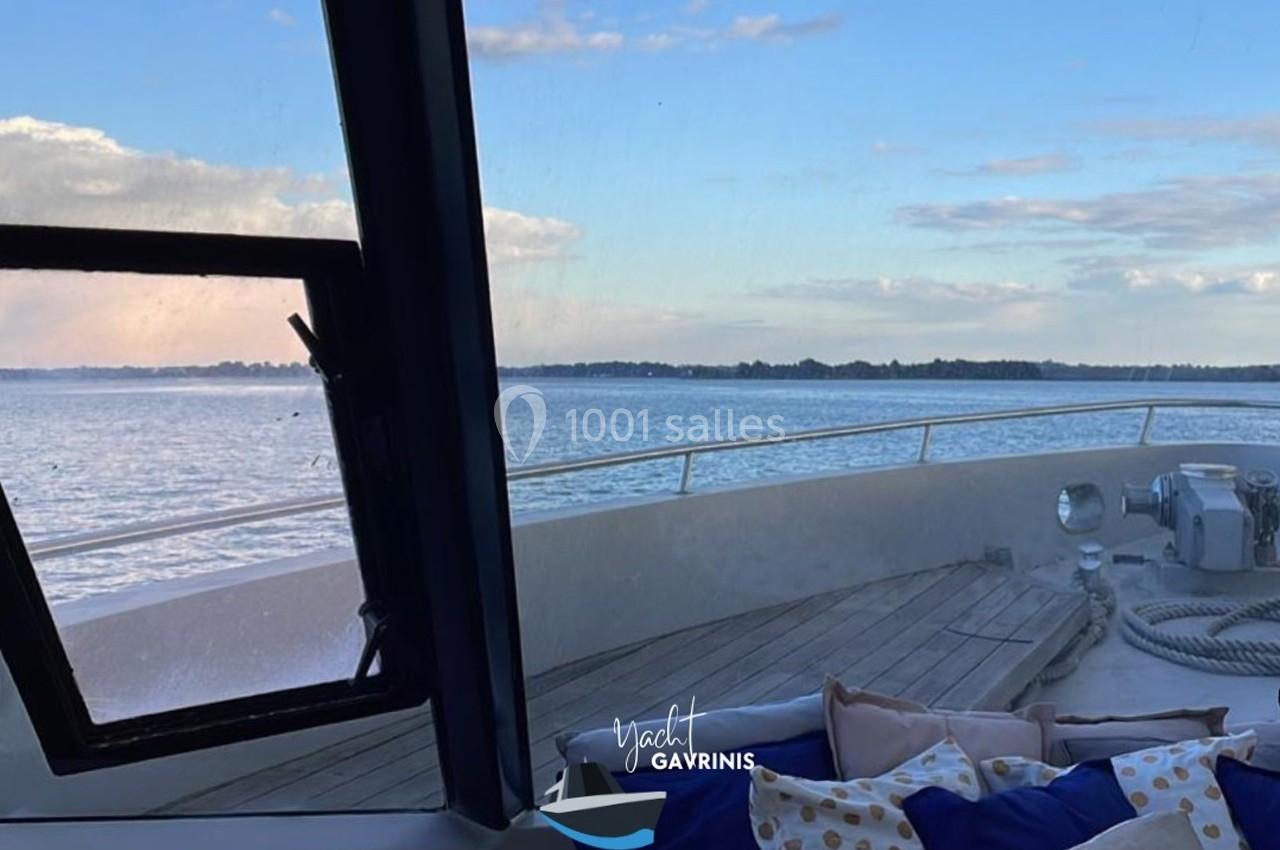 Vue depuis l'intérieur d'un bateau, montrant le pont extérieur, des coussins et un paysage marin sous un ciel dégagé.