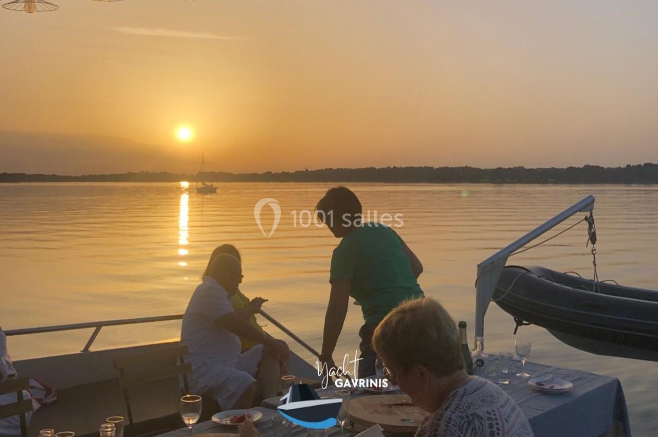 Des personnes dînent sur un bateau au coucher du soleil, avec une vue sur un lac calme et un ciel orangé.