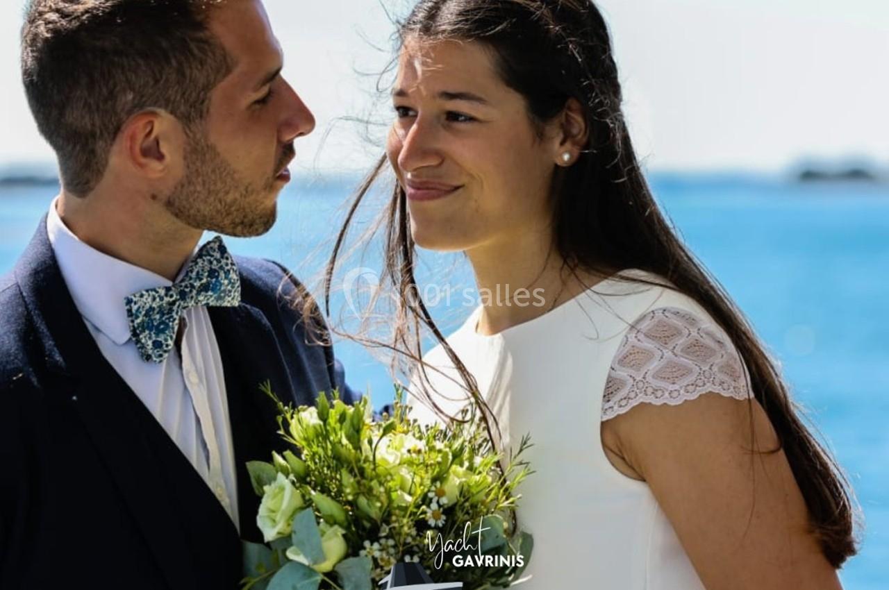 Un couple en tenue de mariage se regarde avec complicité devant un paysage marin.