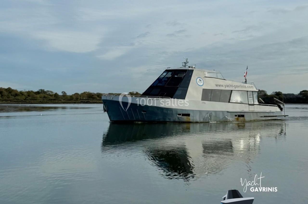 Bateau moderne gris flottant sur une eau calme, avec des arbres en arrière-plan sous un ciel légèrement nuageux.