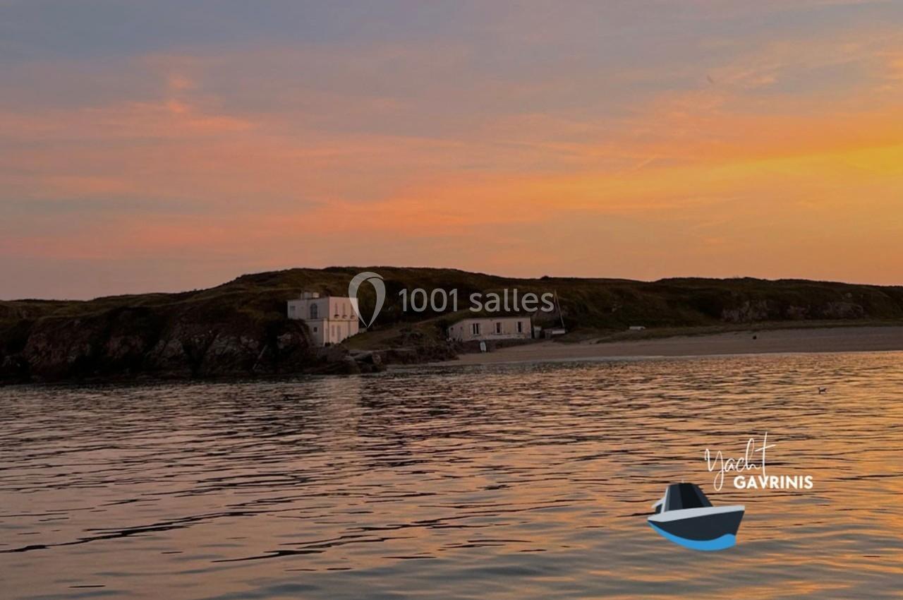 Vue d'une côte rocheuse avec des bâtiments blancs au coucher du soleil, reflétée sur une mer calme.