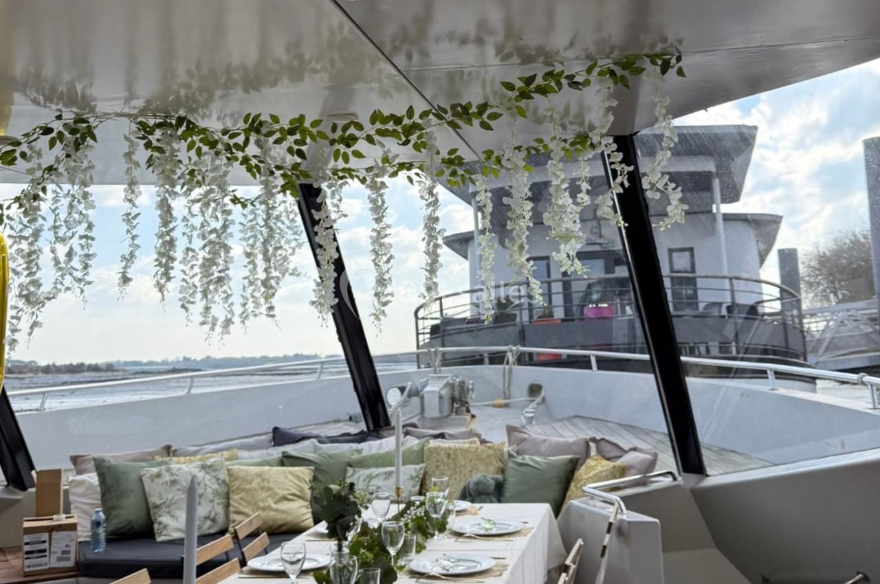 Table élégamment dressée avec des chandeliers et des plantes suspendues, sur un bateau avec vue sur l'eau et des bâtiments…