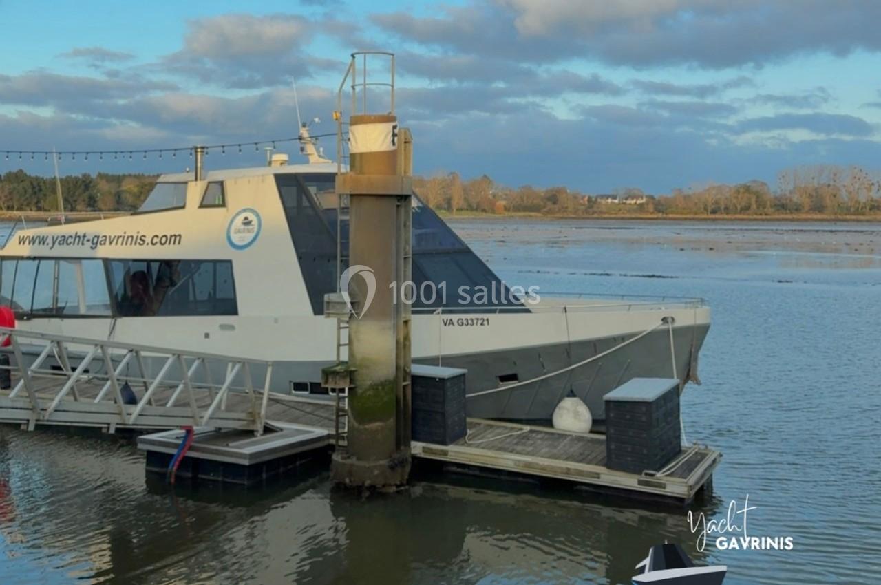Bateau blanc amarré à un ponton sur une rivière, avec un paysage naturel en arrière-plan.