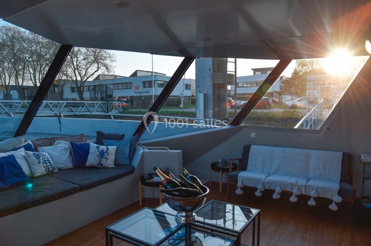 Intérieur d'un bateau avec salon aménagé, vue sur l'extérieur au coucher du soleil à travers de grandes fenêtres.