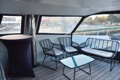 Mobilier en rotin noir avec coussins blancs sur une terrasse en bois couverte, vue sur l'extérieur urbain.