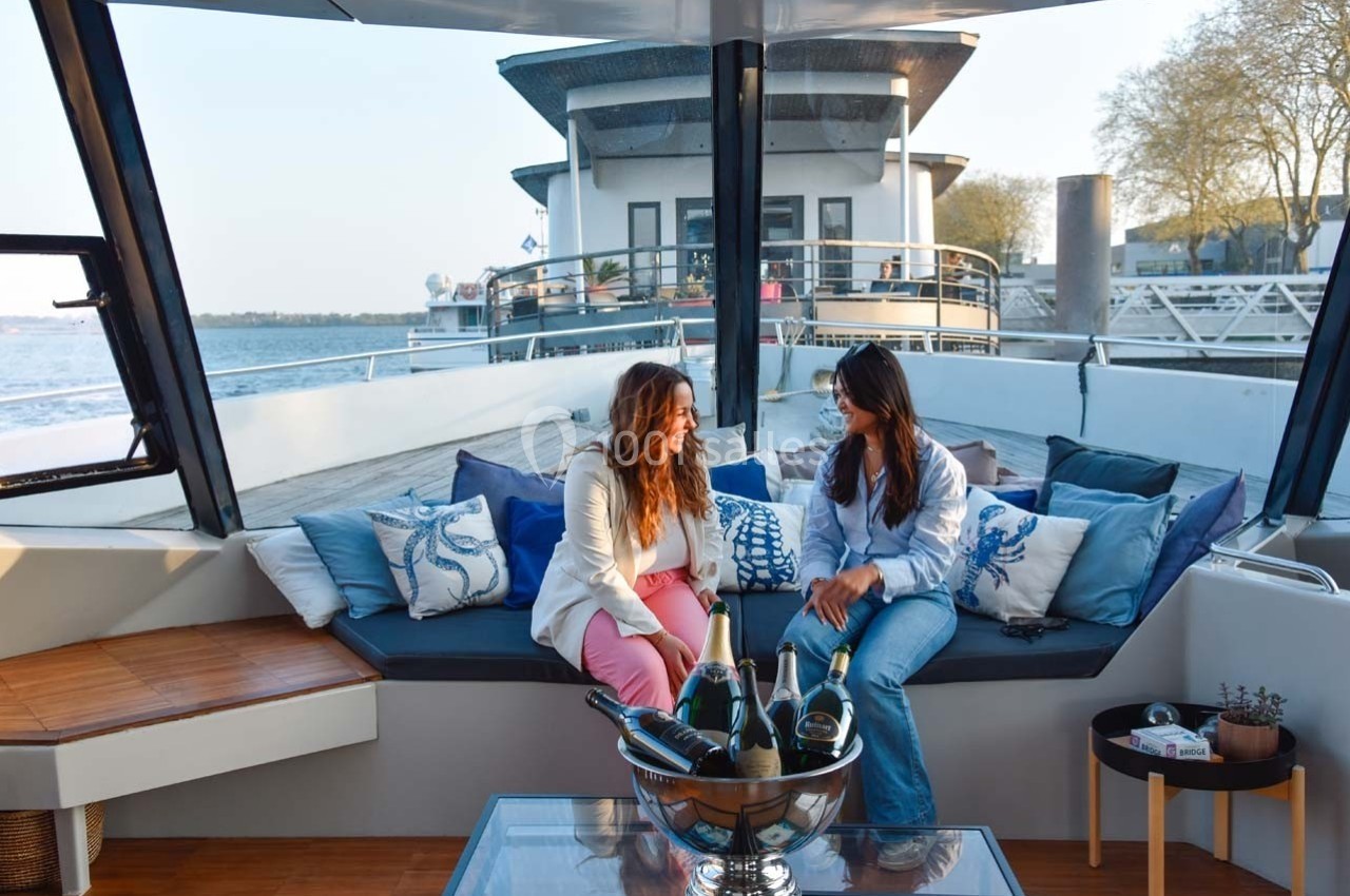 Deux femmes discutent sur un canapé extérieur d'un bateau, entourées de coussins et d'une table avec des bouteilles.