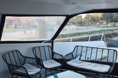 Mobilier en rotin noir avec coussins blancs sur une terrasse en bois couverte, vue sur l'extérieur urbain.