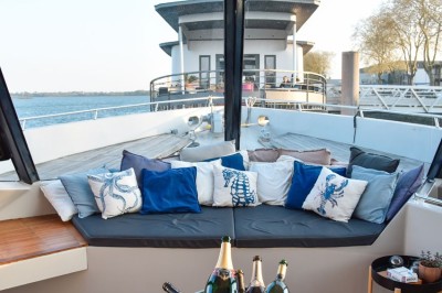 Mobilier en rotin noir avec coussins blancs sur une terrasse en bois couverte, vue sur l'extérieur urbain.