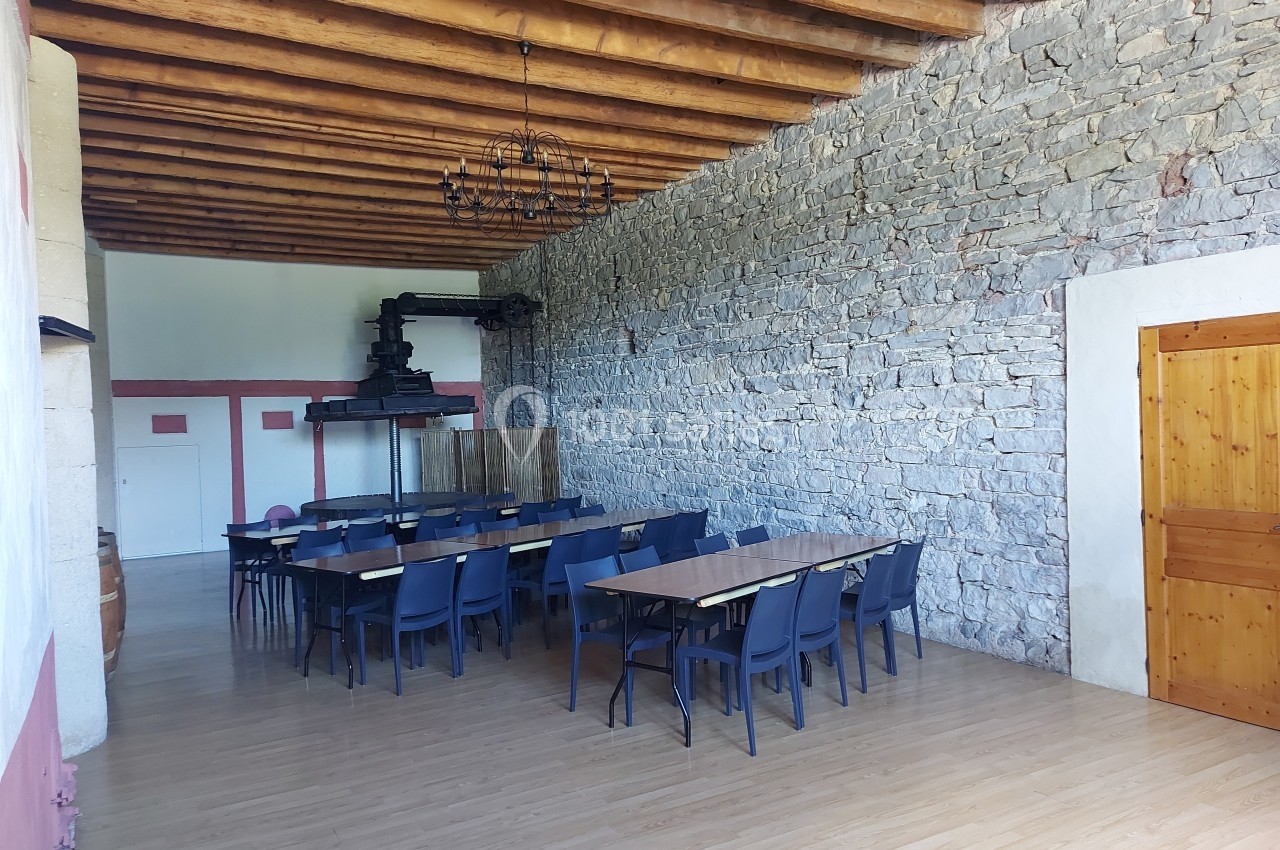 Salle avec des tables et chaises alignées, murs en pierre, plafond en bois avec poutres apparentes et un ancien pressoir.