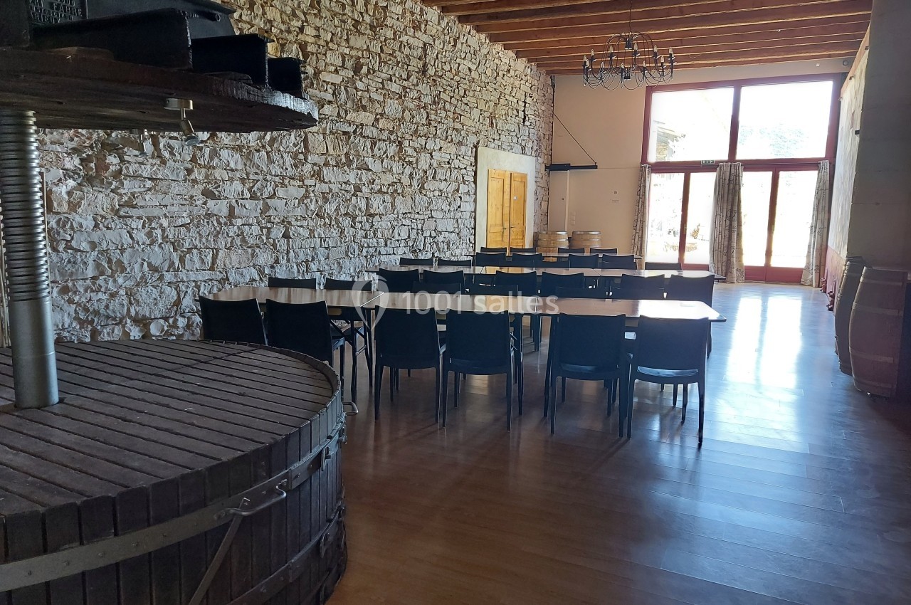Salle avec murs en pierre, tables alignées avec chaises noires, grande baie vitrée et ancien pressoir en bois au premier…
