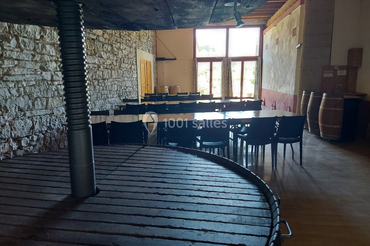 Salle lumineuse avec murs en pierre, tables et chaises noires, tonneaux en bois et grande presse au premier plan.