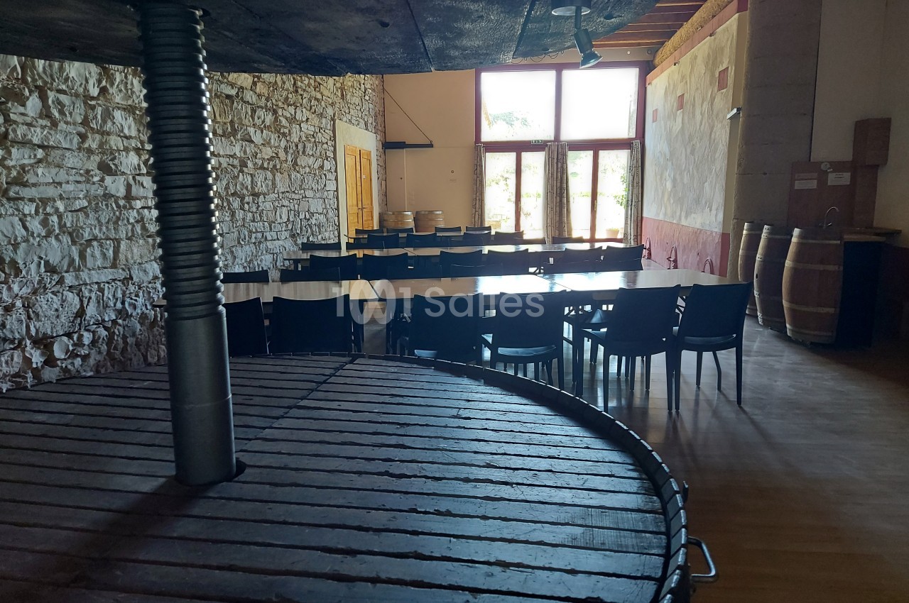Salle lumineuse avec murs en pierre, tables et chaises noires, tonneaux en bois et grande presse au premier plan.