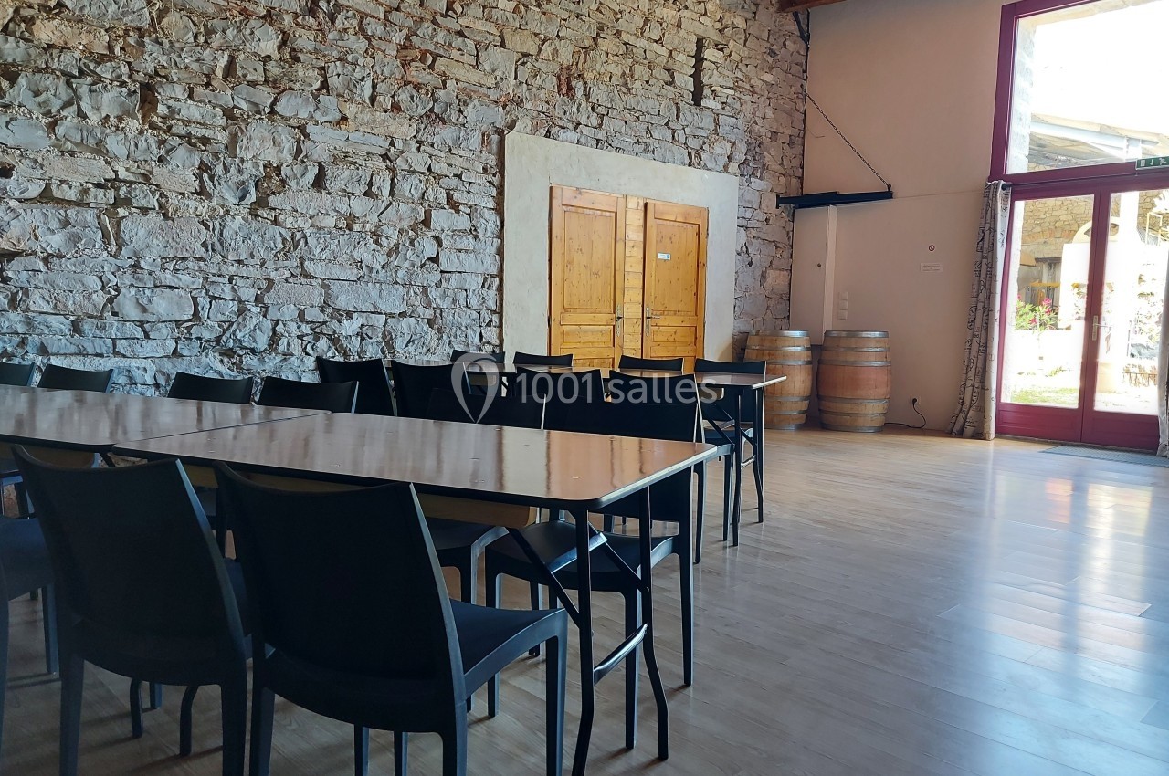 Salle lumineuse avec murs en pierre, tables et chaises noires, barriques en bois et grande porte vitrée.