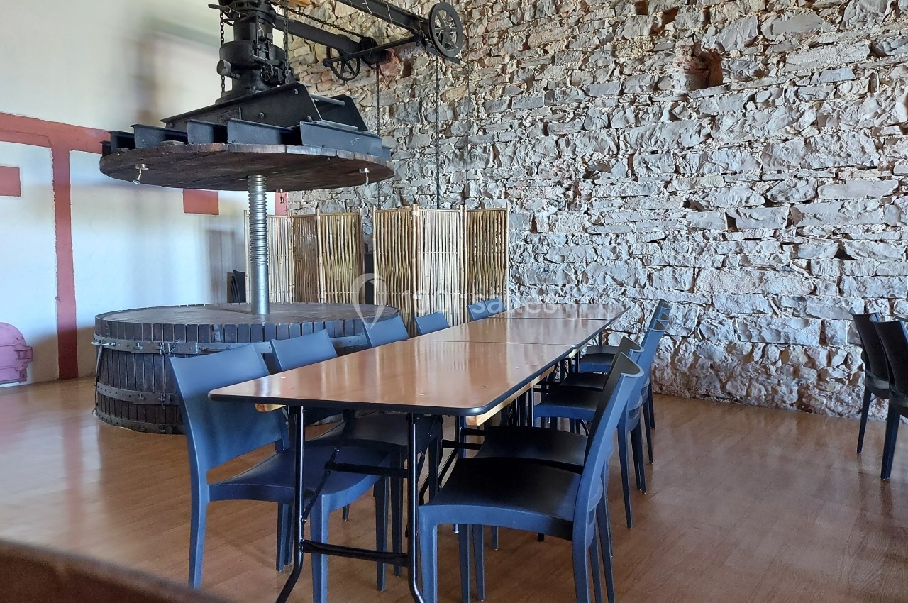 Salle avec une longue table en bois entourée de chaises noires, décorée d'un ancien pressoir et de murs en pierre.