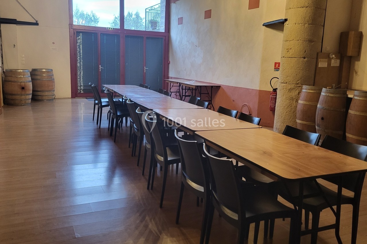 Salle avec une longue table en bois entourée de chaises noires, des tonneaux et un mur en pierre en arrière-plan.