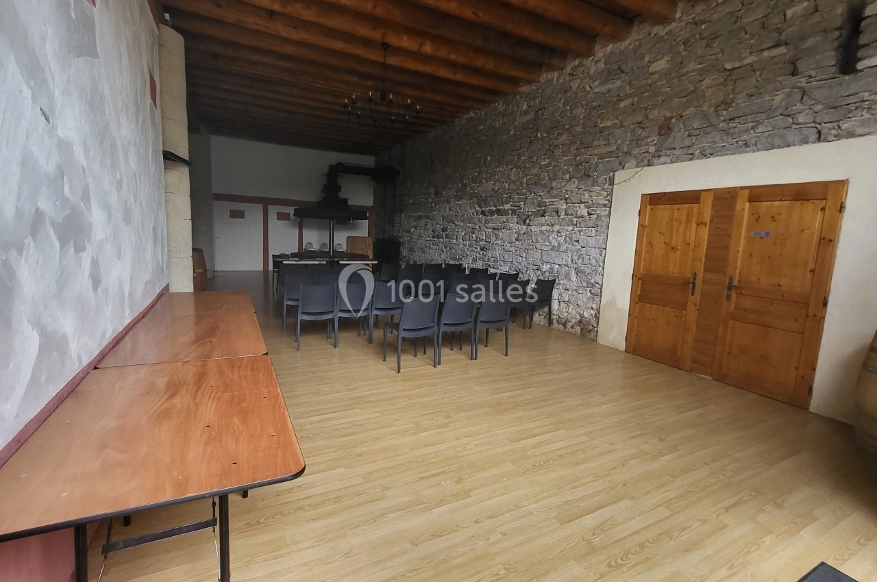 Salle avec murs en pierre et plafond en bois, équipée de chaises noires, tables en bois et tonneaux dans un coin.