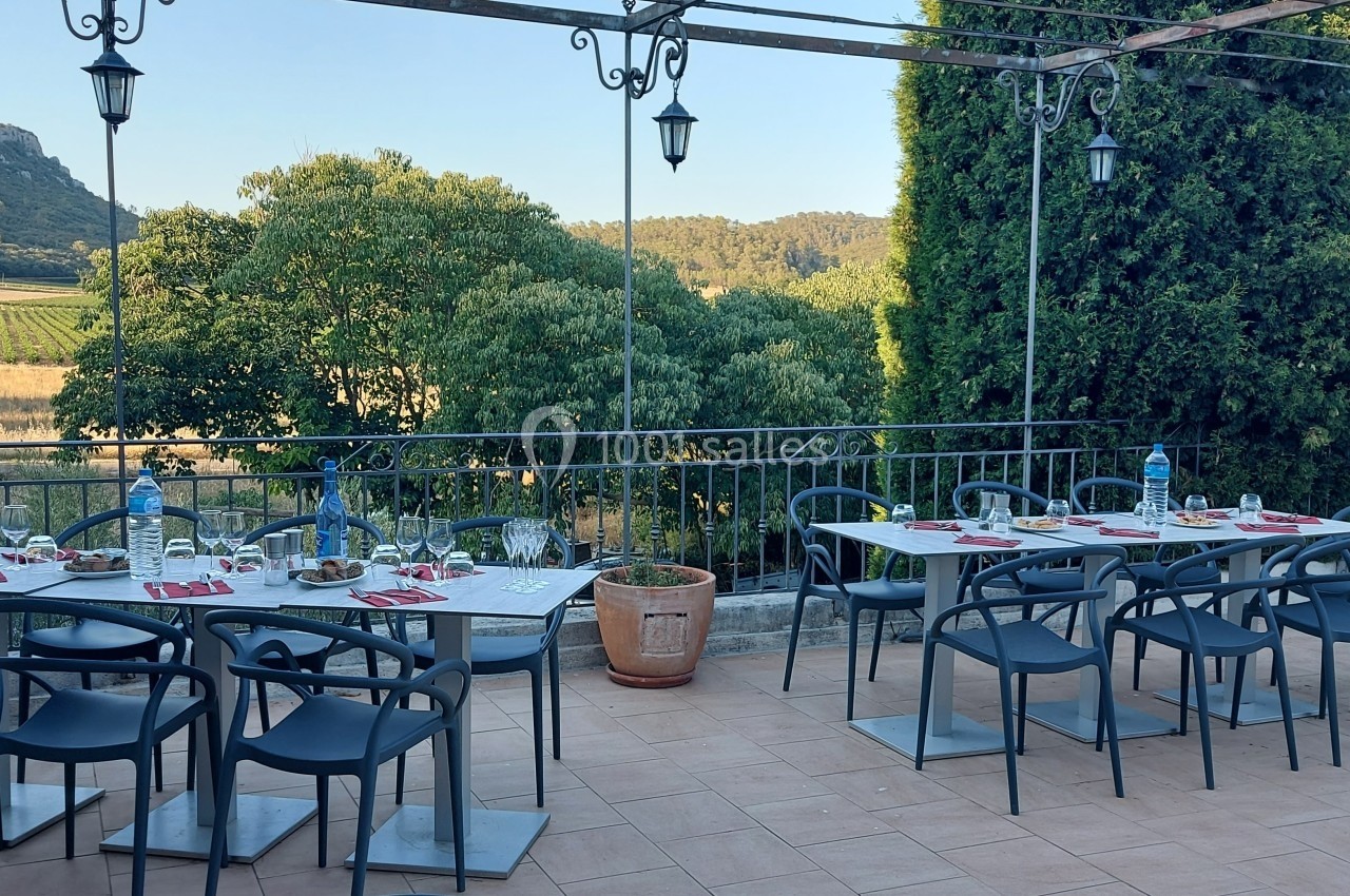 Terrasse extérieure avec tables dressées, chaises noires et vue sur un paysage de campagne avec collines et végétation.