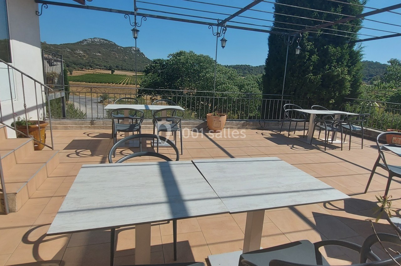 Terrasse ensoleillée avec tables et chaises, entourée de verdure et offrant une vue sur des collines et des vignes.