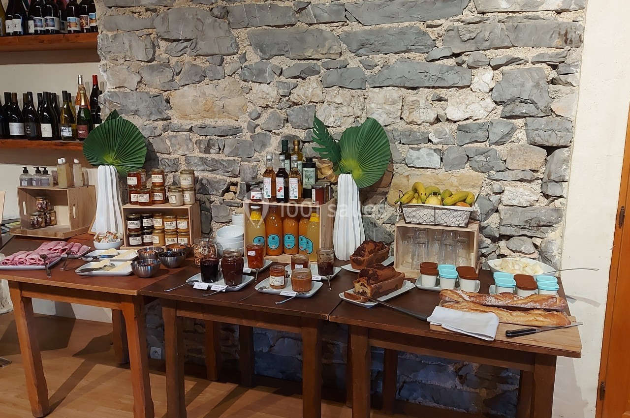 Buffet avec confitures, jus, viennoiseries, charcuteries et fruits, disposé sur des tables en bois devant un mur en pierre.