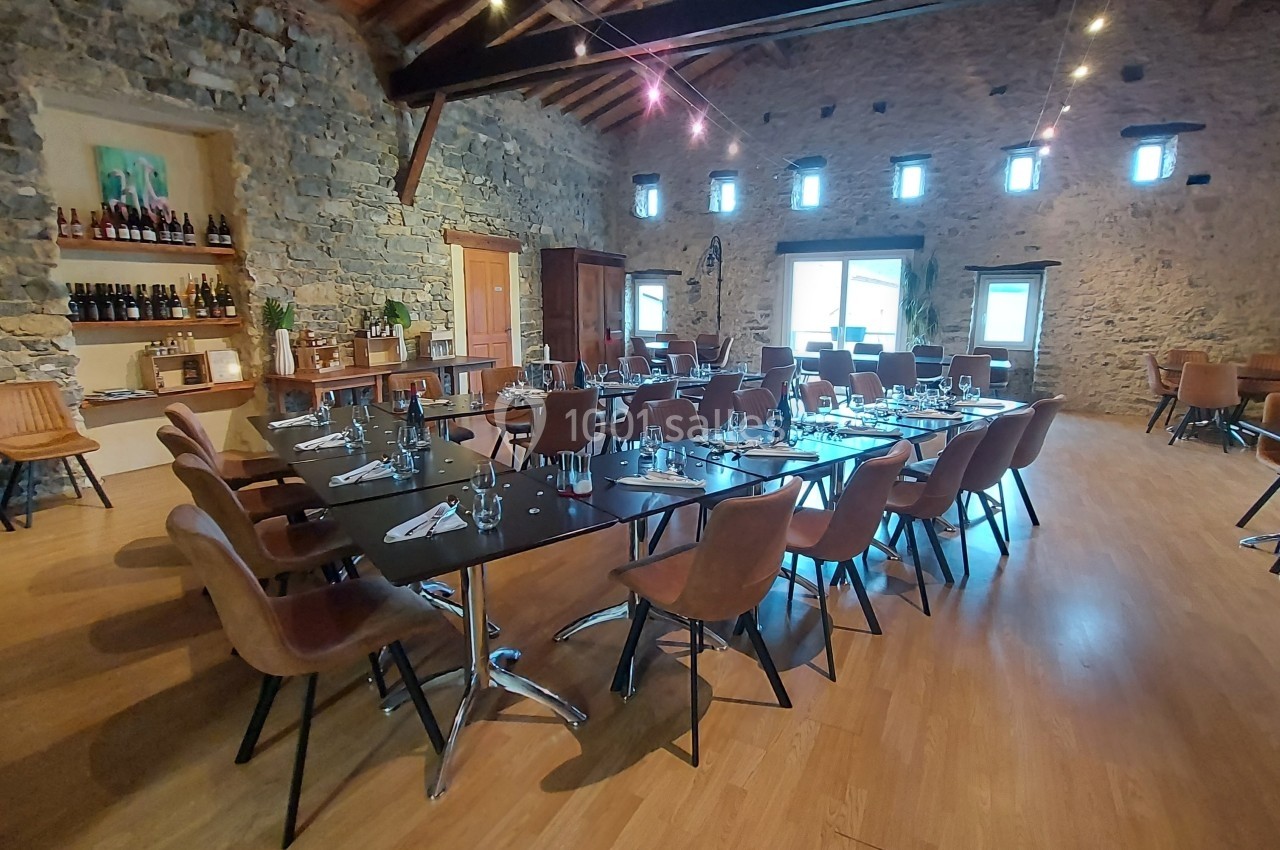Salle en pierre avec tables dressées, chaises en cuir, étagères de bouteilles et lumière naturelle provenant des fenêtres.