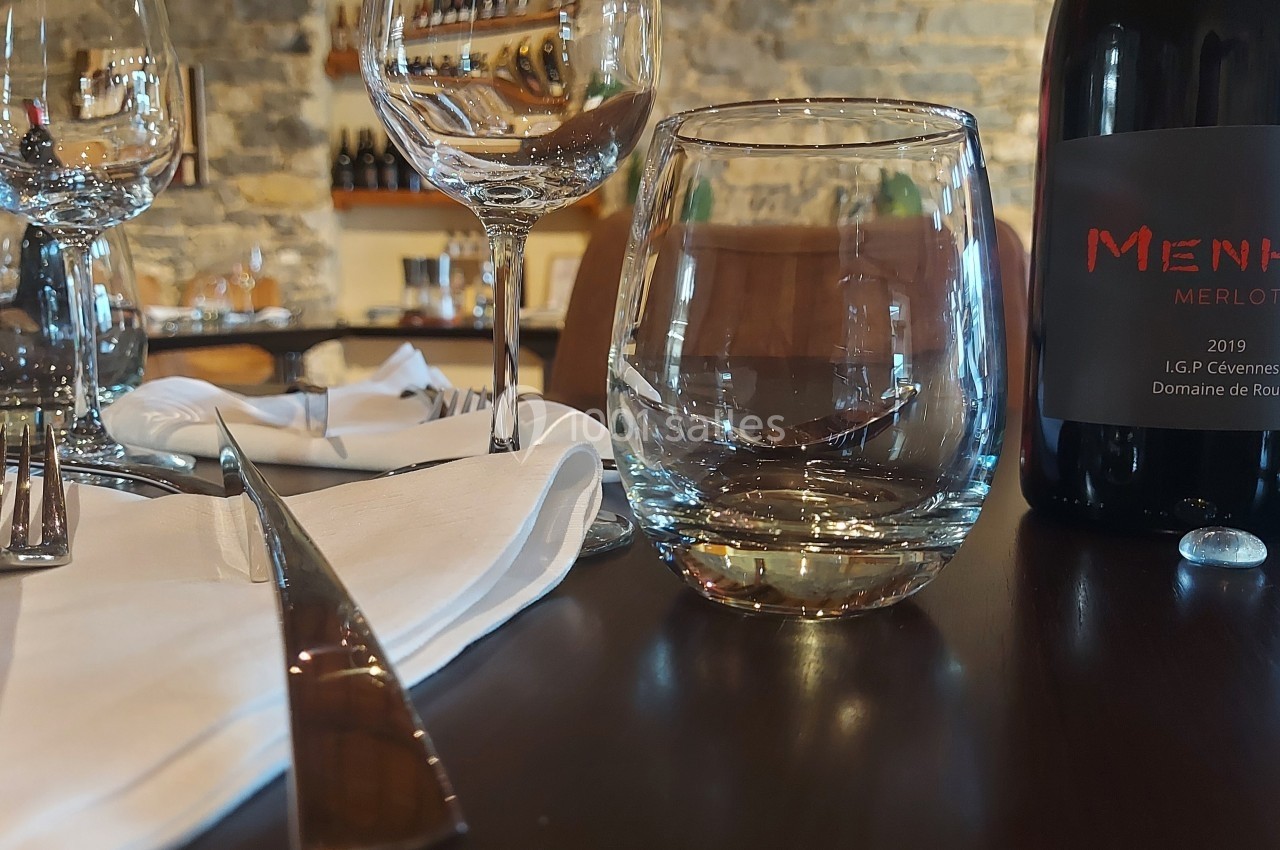 Bouteille de vin rouge, verres et couverts disposés sur une table en bois dans un cadre de restaurant en pierre.