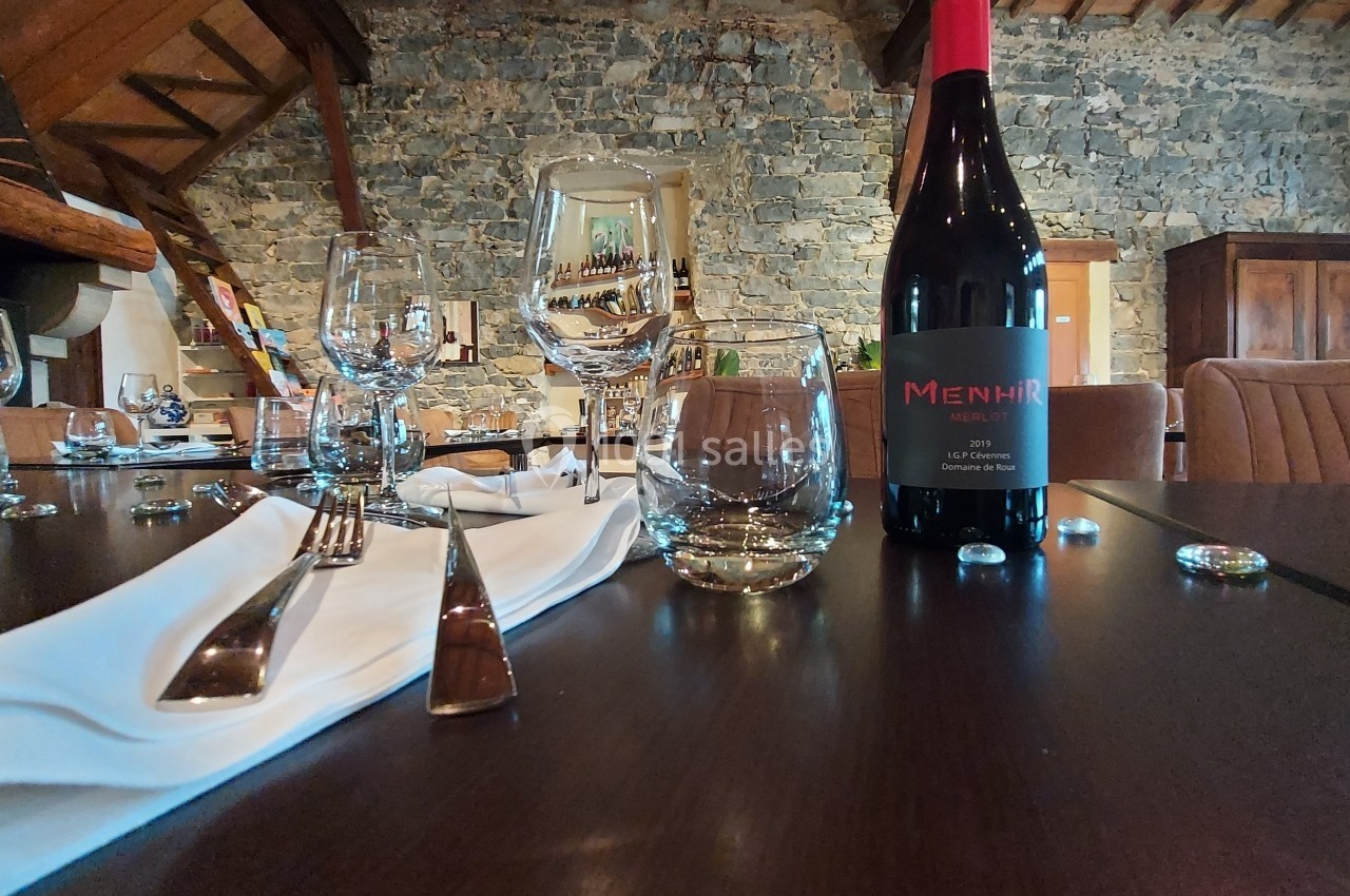 Table dressée avec des verres, couverts et une bouteille de vin rouge dans une salle à murs en pierre apparente.